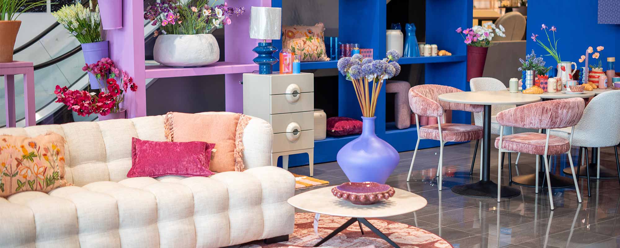 Voorjaarstrend French Flower Market bij Home Center met een crèmekleurige bank, roze fluwelen stoelen en kleurrijke decoraties. Blauwe en paarse accenten, bloemen en speelse accessoires creëren een levendige, romantische sfeer