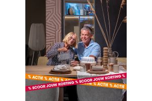Echtpaar zit aan een sfeervol gedekte eettafel in een warme woonomgeving tijdens de pre-opruiming van Kapenga Wonen, met de tekst Scoor jouw korting.