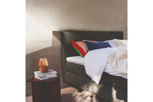 Auping Original boxspring in moderne slaapkamer, met witte dekens, kleurrijke kussens en een nachtkastje met boeken en lamp.