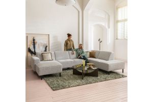 Woonkamer met royale lichtgrijze hoekbank op zwarte poten, geplaatst op een groen vloerkleed met ronde salontafel, terwijl twee personen ontspannen genieten van het interieur.