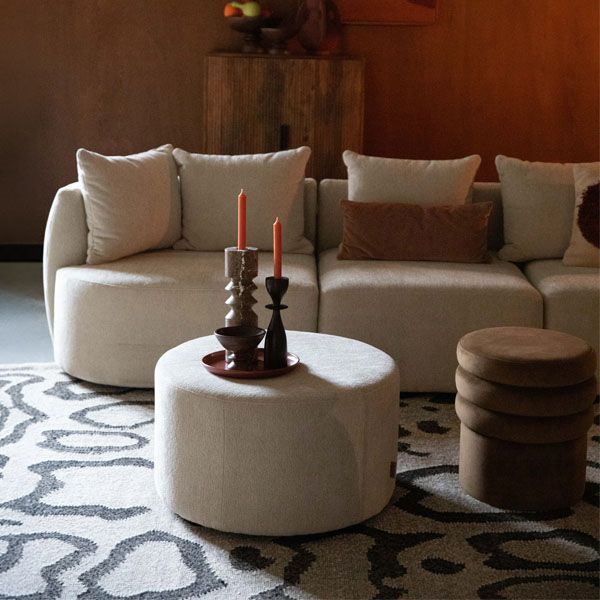 Sfeervolle woonkamer met een beige modulaire bank, ronde poef en houten bijzettafel op een grafisch vloerkleed in warme aardetinten.