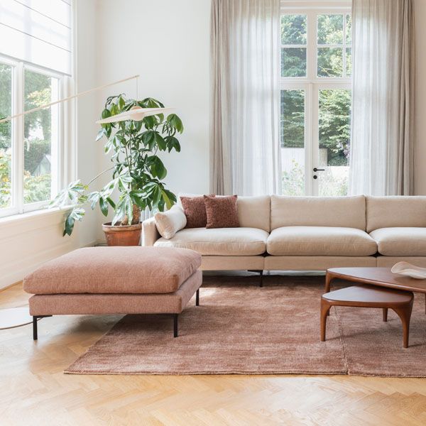 Lichte woonkamer met moderne beige hoekbank, roze ottoman, houten salontafels en groot vloerkleed in warme natuurtinten