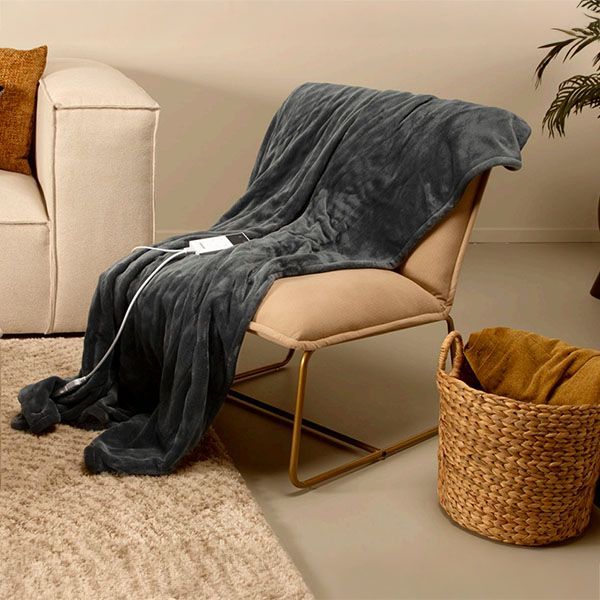 Donkergrijze elektrische fleece deken van Silvana, gedrapeerd over een beige stoel in een warme woonkamer met mand en vloerkleed.