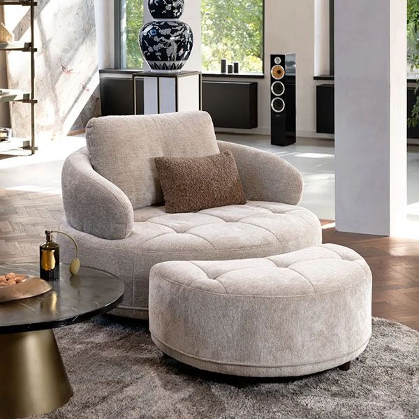 Fauteuil Borgia van Henders & Hazel in een moderne woonkamer, bekleed met zachte beige stof en inclusief bijpassende hocker op een vloerkleed.