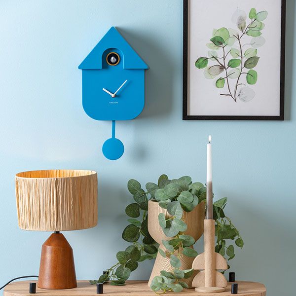 Blauwe Karlsson Modern Cuckoo wandklok aan een lichtblauwe muur, met eronder een houten tafellamp, planten en een kaarshouder.