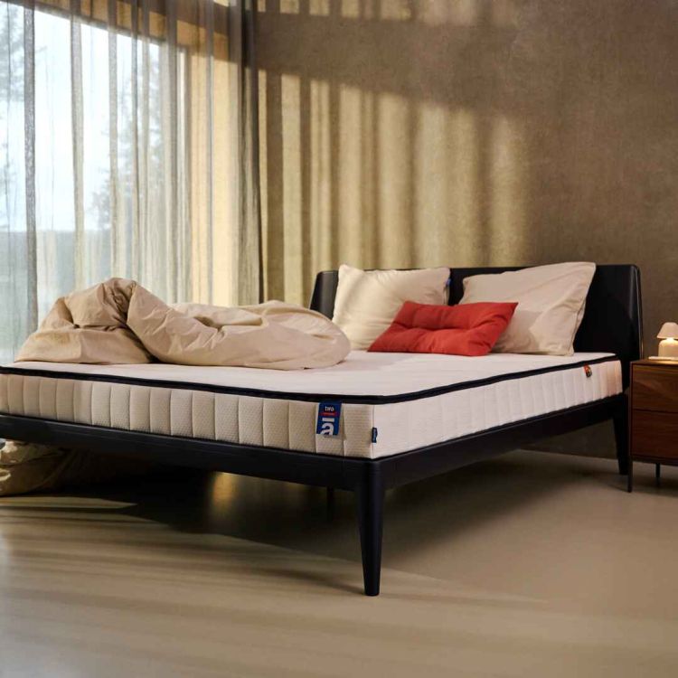 Auping Two matras op een modern zwart bedframe in een sfeervolle slaapkamer met lichte dekbedden, witte kussens en een rood sierkussen.