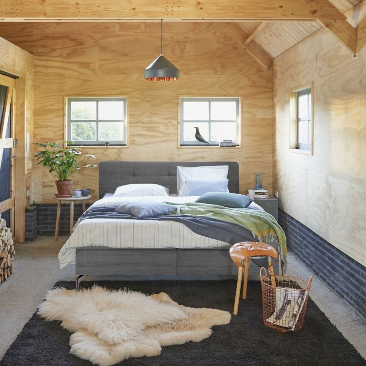 Sfeerfoto van een Auping boxspring in een houten slaapkamer, bekleed met grijze stof, opgemaakt met licht beddengoed, plaid en kussens, met vloerkleed en natuurlijke accessoires.