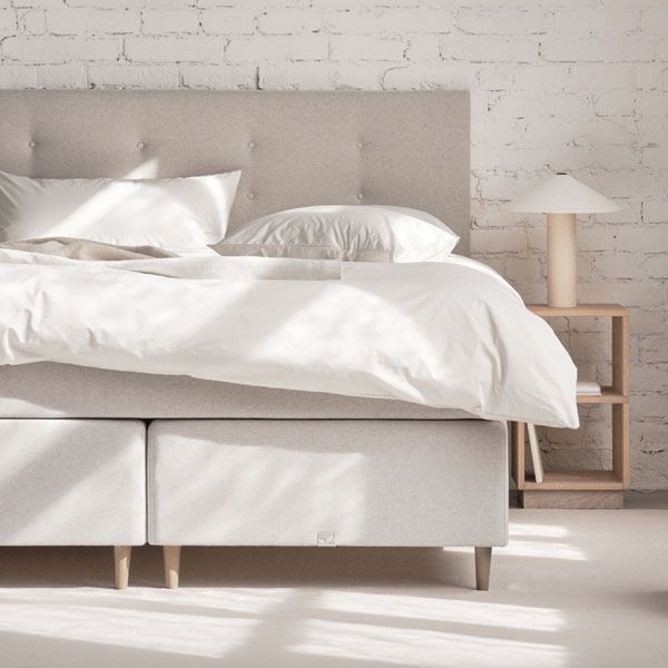 Licht beige boxspring bed met gestoffeerd hoofdbord, witte dekens en kussens in een minimalistische slaapkamer met houten nachtkastje en lamp