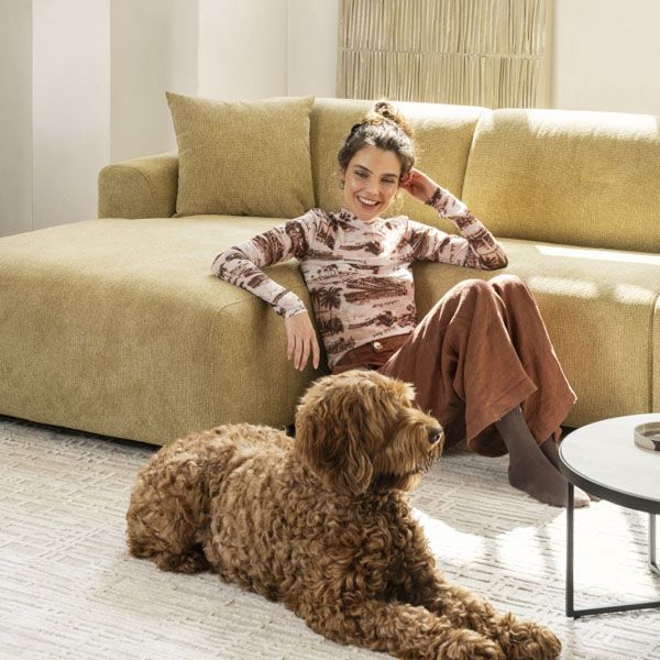 Vrouw ontspant op beige hoekbank met hond op licht vloerkleed in warme, moderne woonkamer