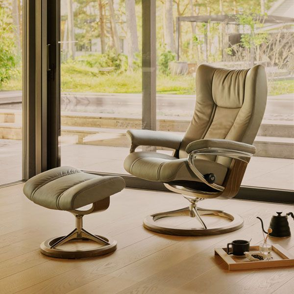 Stressless Wing fauteuil met voetenbank in licht leer, geplaatst bij groot raam in moderne woonkamer met natuurlijke lichtinval.