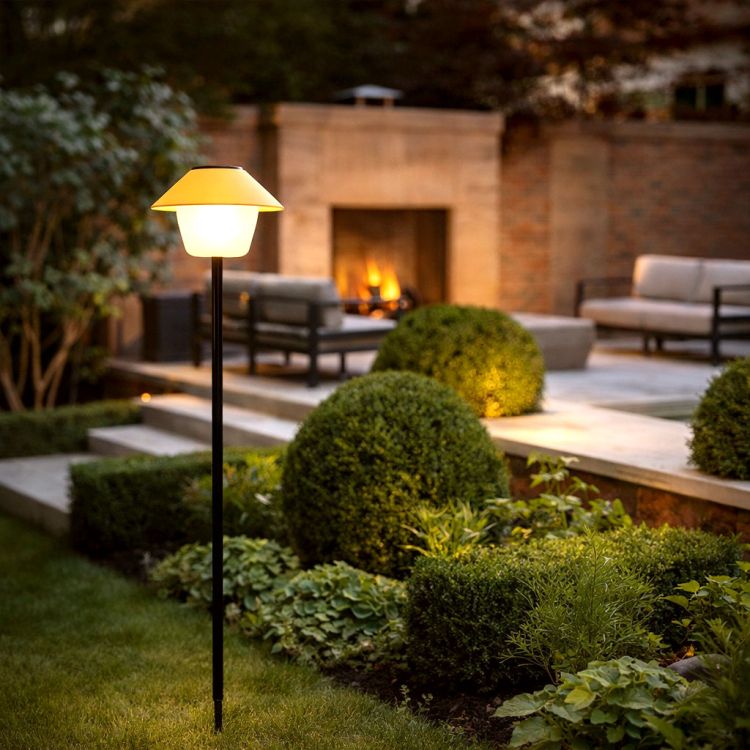 Moderne tuinlamp op een slanke zwarte paal die warm licht geeft langs een tuinpad met strak gesnoeide struiken en een terras met buitenhaard op de achtergrond in de schemering.