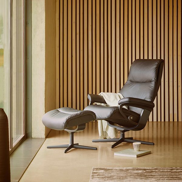 Stressless relaxfauteuil met bijpassende voetenbank in donker leer, geplaatst in een warme houten setting met plaid en koffiekopje.