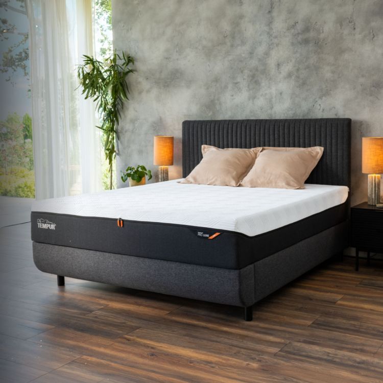 TEMPUR boxspring met donkergrijs hoofdbord en matras in een moderne slaapkamer met houten vloer, sfeerverlichting en grote raampartijen.