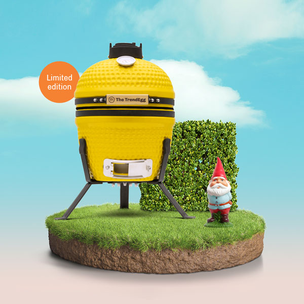 Limited edition gele TrendEgg kamado barbecue op grasplateau met buxushaag, tuinkabouter en blauwe lucht op de achtergrond.