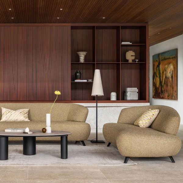 Woonkamer met twee beige banken en een ronde salontafel, geplaatst voor een wandkast in donker hout, met vloerlamp, accessoires en kunst aan de muur.