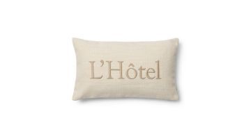 Close-up van de Rivièra Maison kussenhoes in crèmekleur met de tekst ‘L’Hôtel’ in reliëf geborduurd op de voorkant, tegen een witte achtergrond.