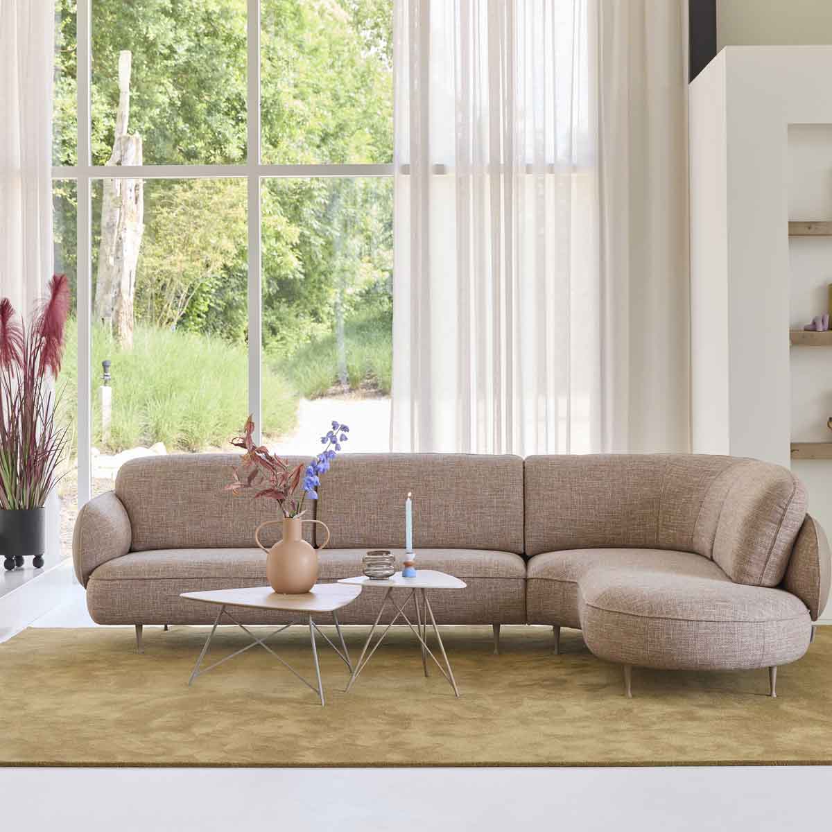 Beige hoekbank van Bert Plantagie met afgeronde vormen in lichte woonkamer met hoge ramen, zacht vloerkleed en moderne salontafel.