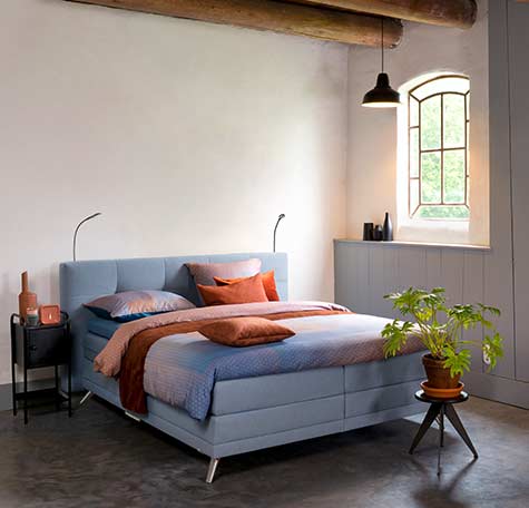 Lichtblauwe boxspring van Avek met roestkleurig beddengoed in sfeervolle slaapkamer met houten balken en industrieel hanglampje