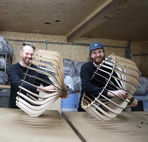 Twee lachende mannen van Blij design tonen houten lamellenframes van designlampen in een ambachtelijke werkplaats met houten plafond.