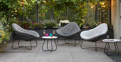 Drie luxe loungestoelen met metalen onderstel en grijze kussens, gecombineerd met bijzettafels in een sfeervolle tuin
