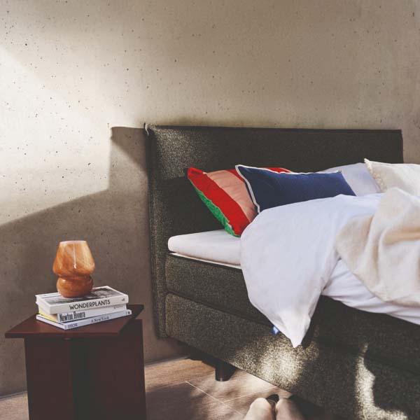 Auping Original boxspring in moderne slaapkamer, met witte dekens, kleurrijke kussens en een nachtkastje met boeken en lamp.