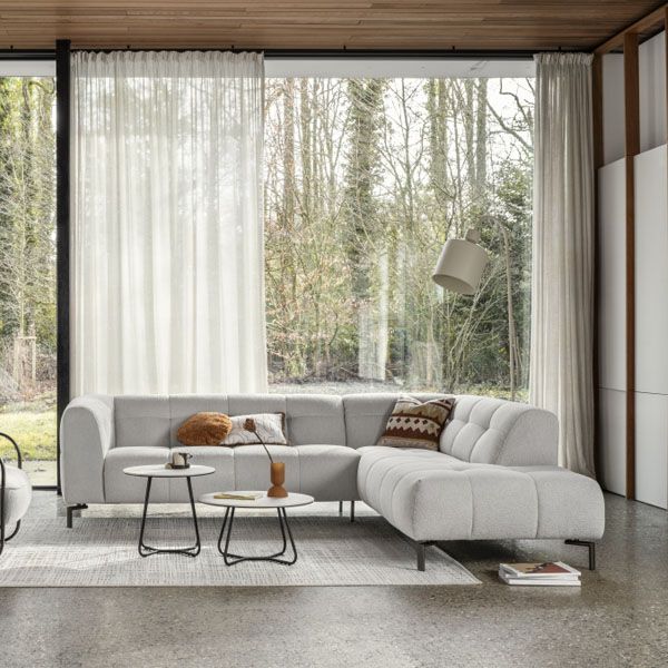 Lichtgrijze hoekbank met afgeronde vormen in moderne woonkamer, voor groot raam met witte gordijnen en twee ronde bijzettafels.
