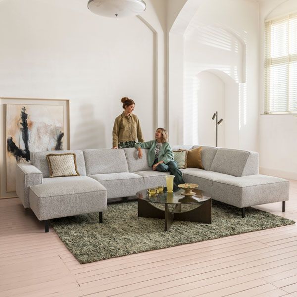 Woonkamer met royale lichtgrijze hoekbank op zwarte poten, geplaatst op een groen vloerkleed met ronde salontafel, terwijl twee personen ontspannen genieten van het interieur.