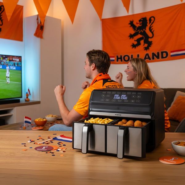 Twee supporters in oranje juichen voor de televisie tijdens een voetbalwedstrijd, met een airfryer op tafel als onderdeel van de WK-actie van Home Center en Kapenga Wonen.