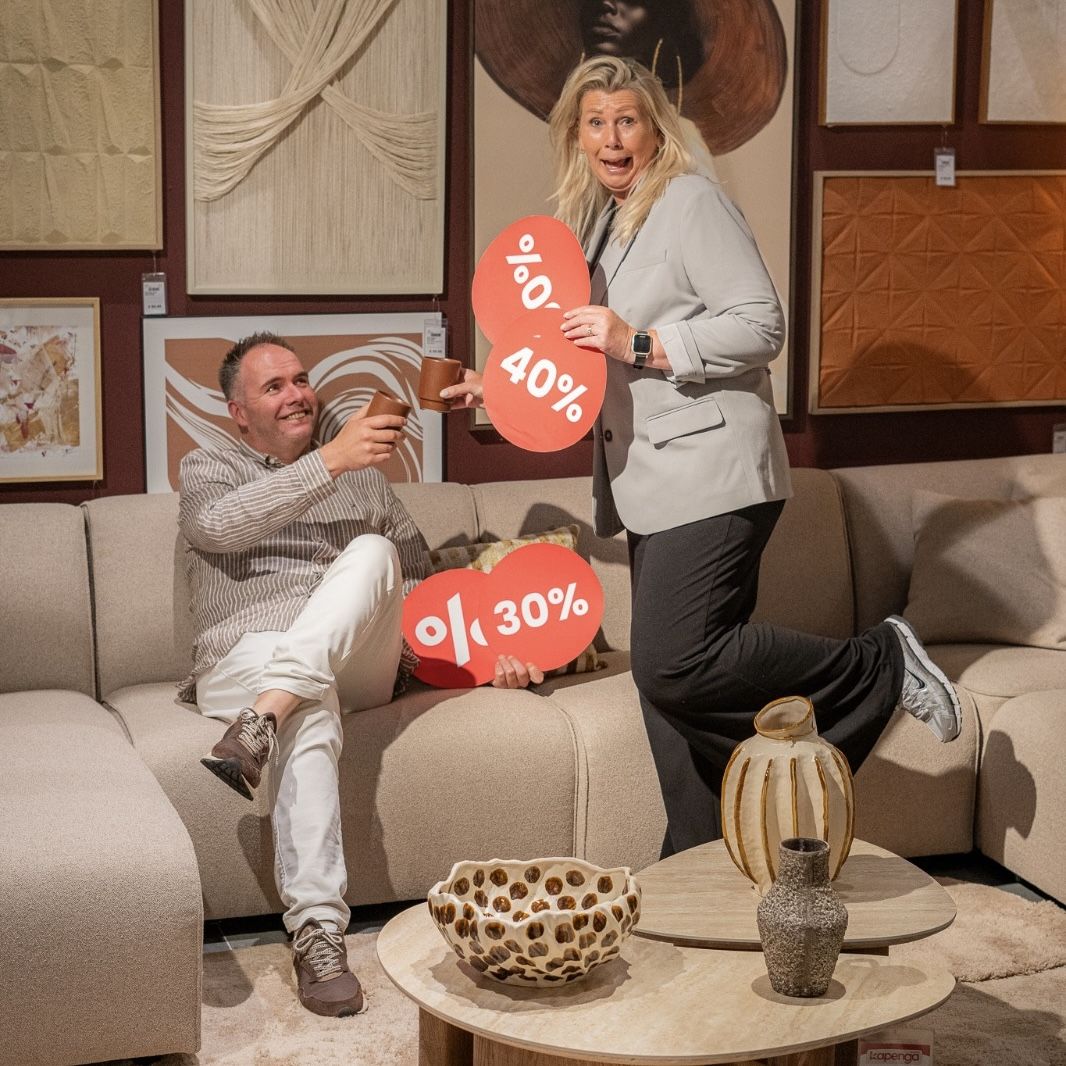 Pre-Opruiming bij Kapenga Wonen met aantrekkelijke kortingen Andre en Angelique van Kapenga Wonen promoten de Pre-Opruiming met kortingsborden in een sfeervolle woonkameropstelling.
