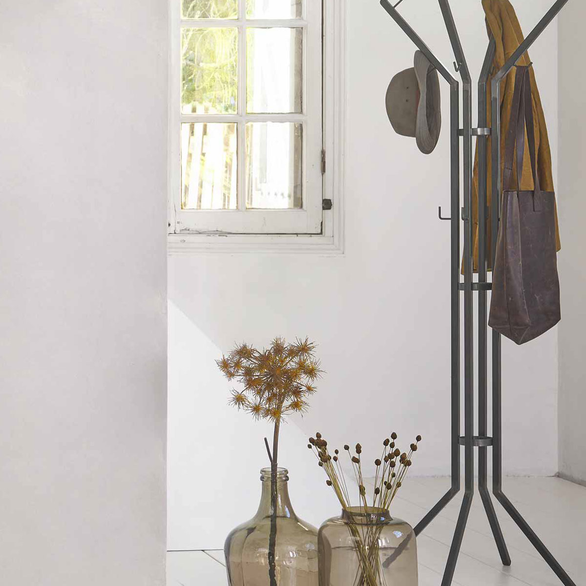Minimalistische metalen kapstok in een lichte hal, voorzien van jas, hoed en tas. De ruimte is gestyled met transparante vazen en gedroogde takken.