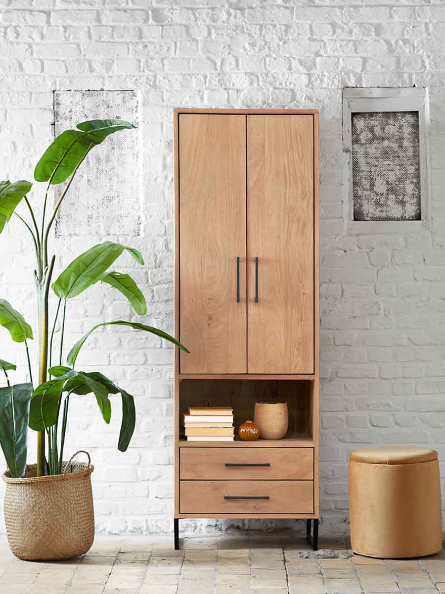 Hoge houten kast met twee deuren, open vak en lades, afgewerkt met zwarte metalen grepen, tegen een witte bakstenen muur met decoratieve accessoires en kamerplant.