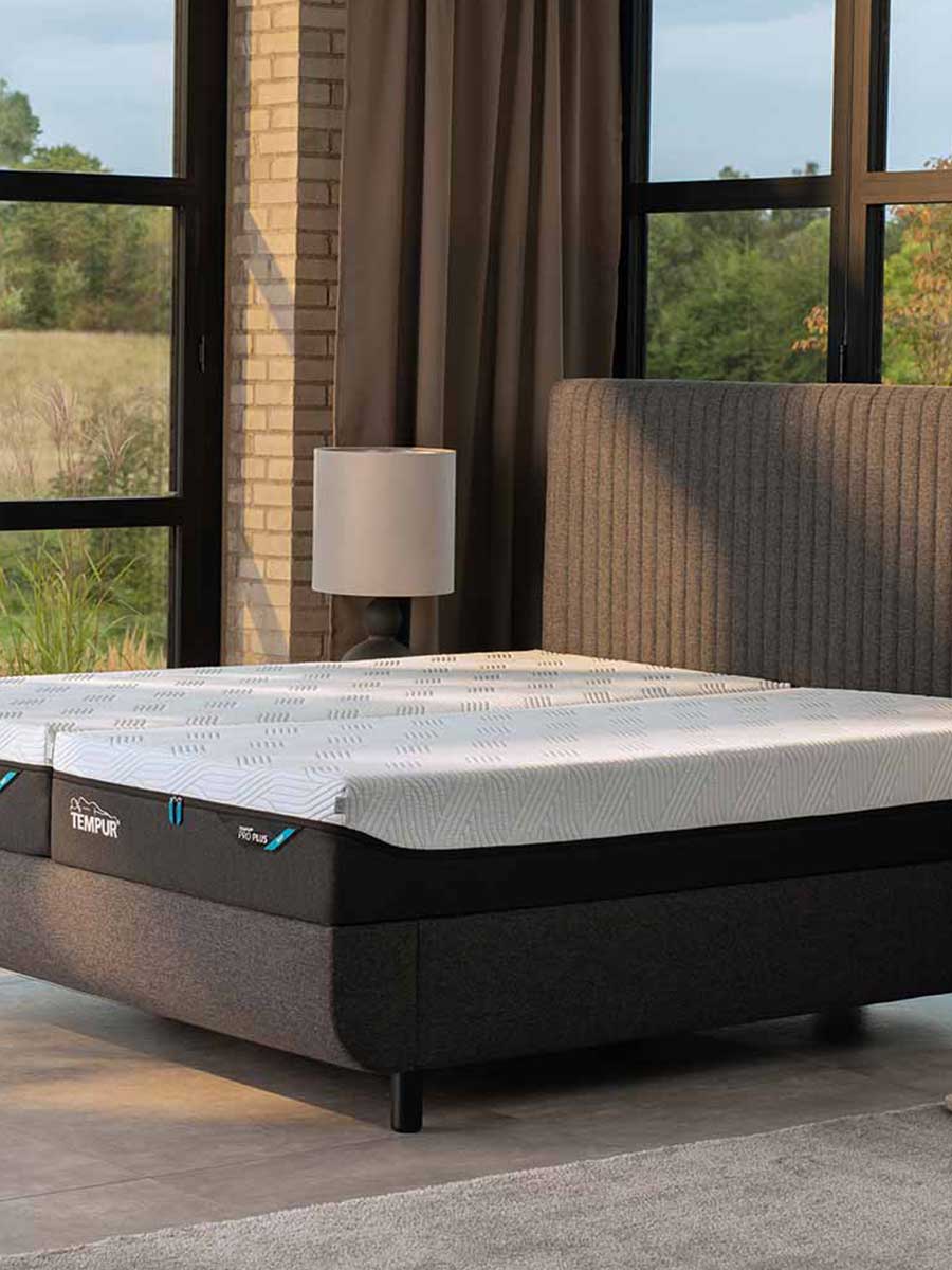 Donkere boxspring met Tempur-matras en geribbeld hoofdbord, geplaatst in een moderne slaapkamer met grote ramen en uitzicht op het landschap.