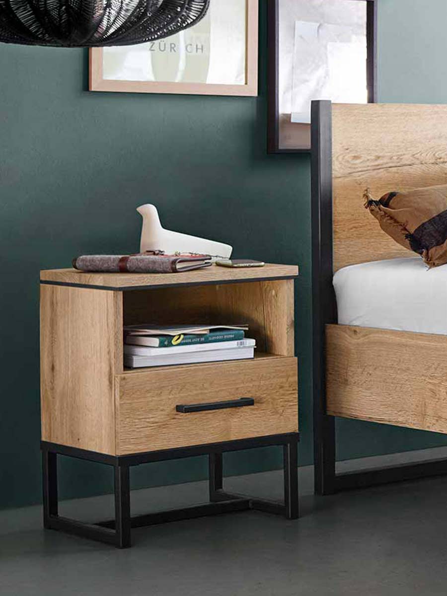 Houten nachtkastje met lade en open vak op zwart metalen frame, geplaatst naast een bed met houten hoofdbord in een modern industrieel interieur.