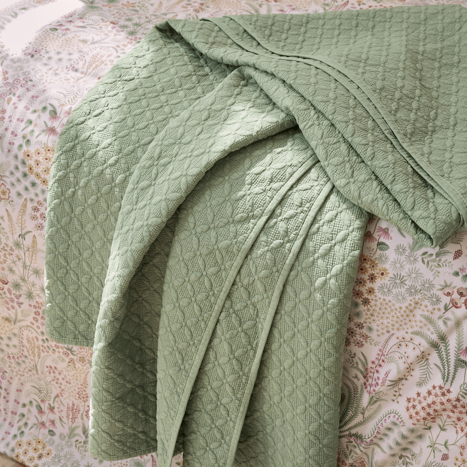 Groene gewatteerde plaid met bloemstructuur gedrapeerd over een bed met bloemenprint beddengoed.