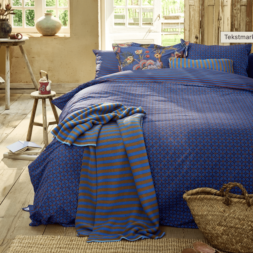 Blauw dekbedovertrek met grafisch patroon, sierkussens en plaid in een sfeervol ingerichte slaapkamer.