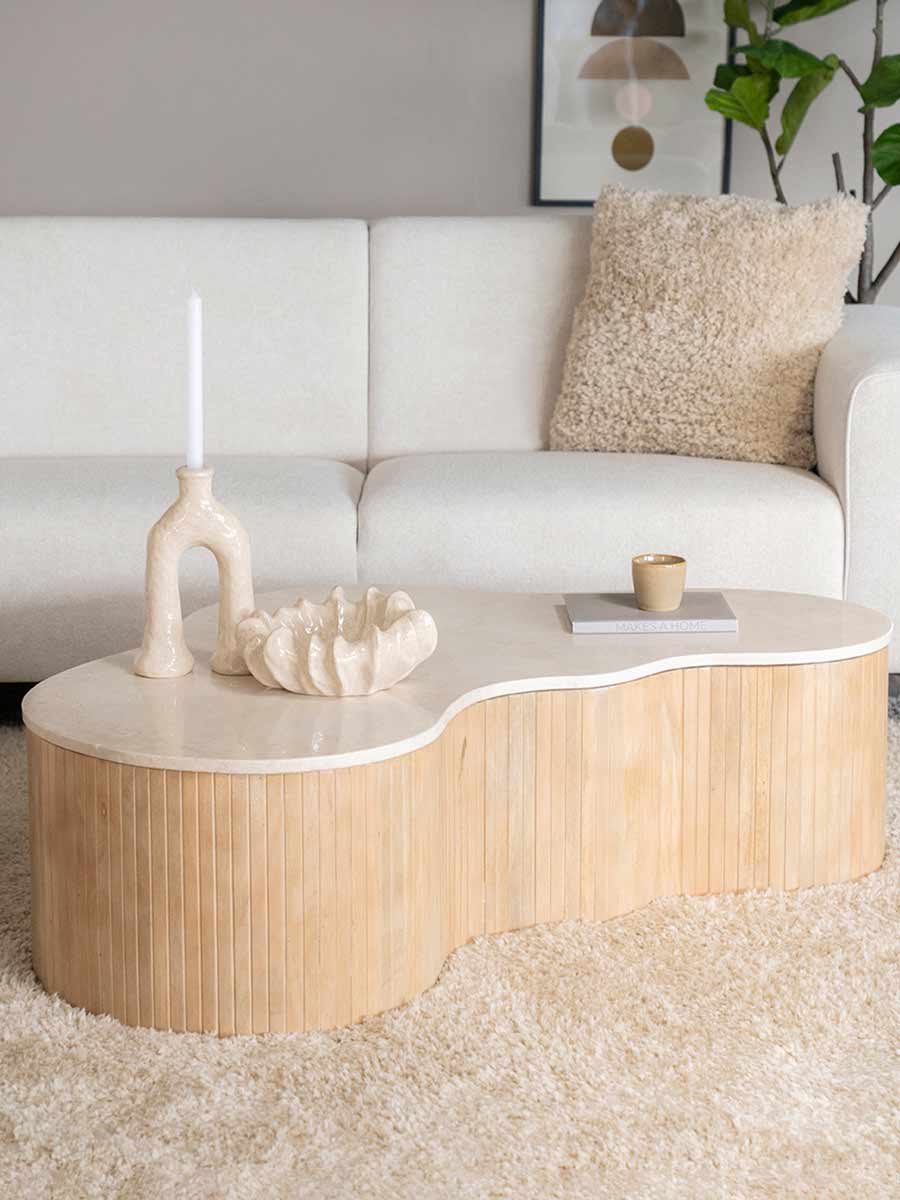 Organisch gevormde houten salontafel met lichte afwerking en verticale latstructuur, geplaatst op een hoogpolig vloerkleed in een modern ingerichte woonkamer.
