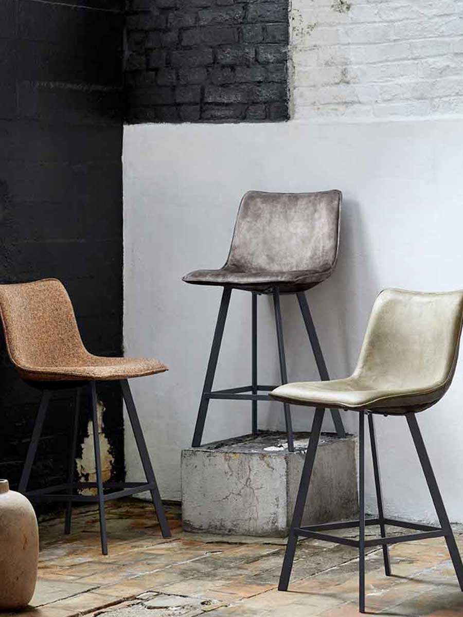 Drie industriële barkrukken met stoffen en leren bekleding in warme tinten, op zwarte metalen poten, gestyled tegen een zwart-witte muur met beton- en baksteenelementen.