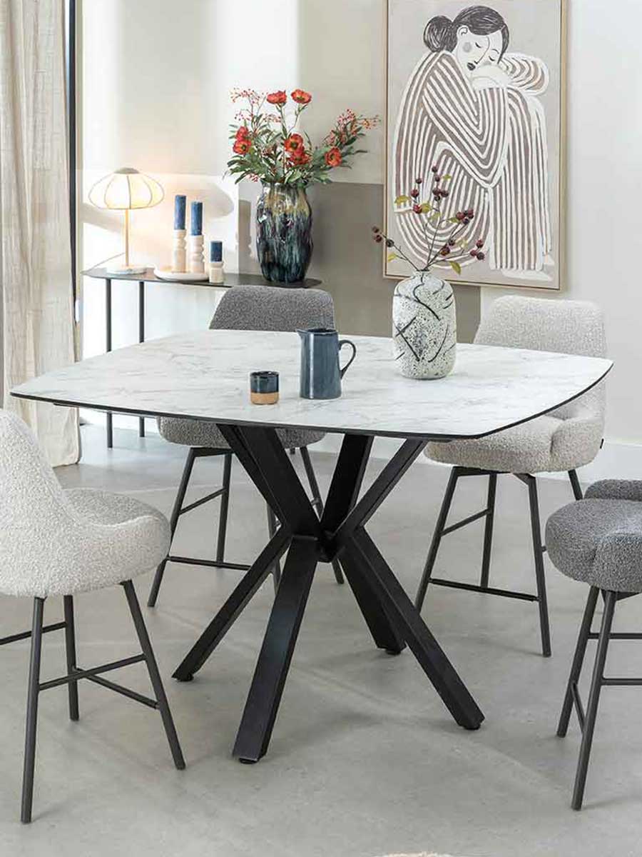 Stijlvolle eetopstelling met vier licht gemêleerde barkrukken en een vierkante bartafel met marmerlook blad en zwart metalen kruispoot, in een licht interieur met kunst en accessoires.