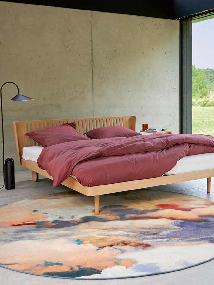 Minimalistisch houten bed met lattenhoofdbord, op een ronde kleurrijke vloerkleed met roze dekbedovertrek, in een slaapkamer met betonnen wand en groot raam.