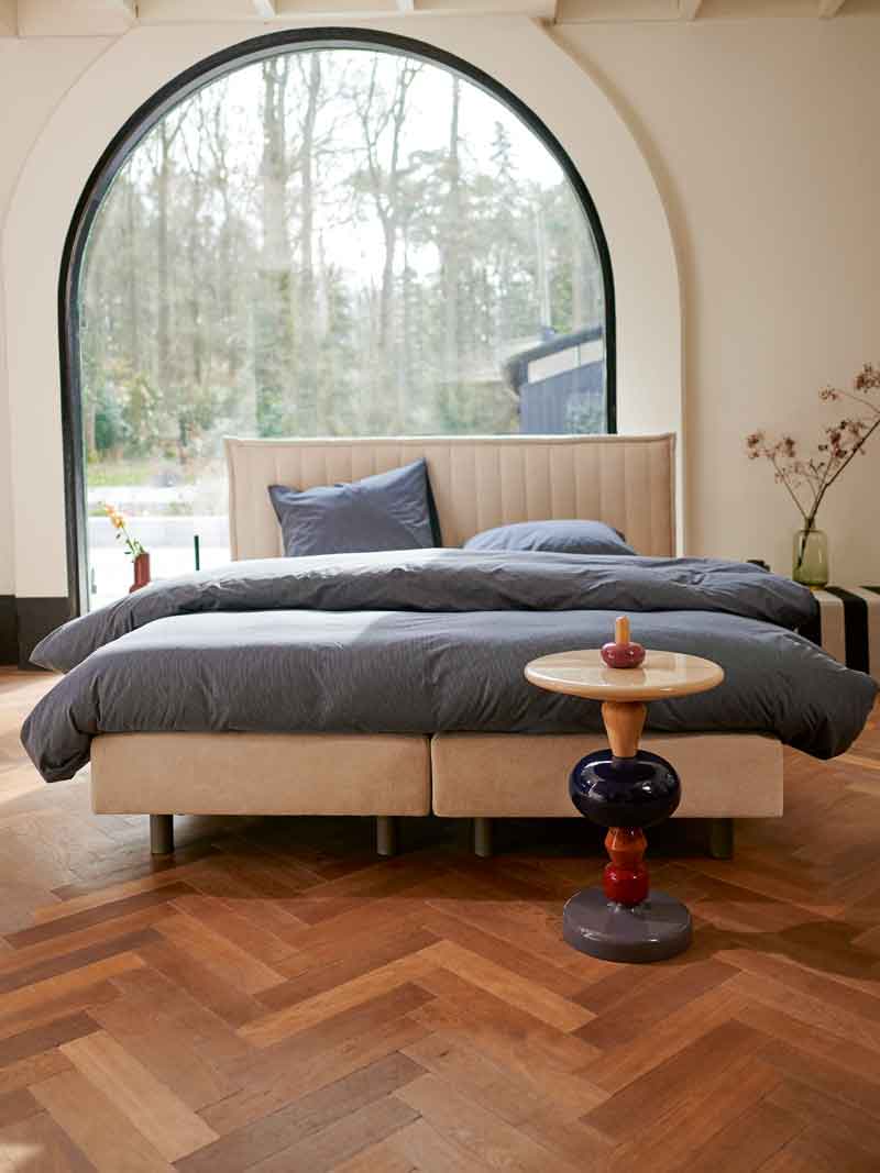 Grijze boxspring met strak hoofdbord in een warme houten slaapkamer. Het bed is aangekleed met wit beddengoed, een groene plaid en een schapenvacht op een donker vloerkleed.