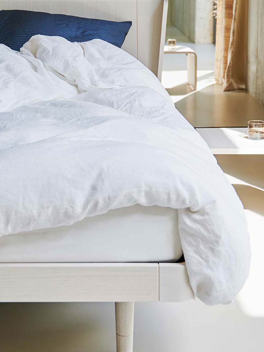 Detailopname van een wit donzig dekbed op een licht houten bedframe, in een moderne slaapkamer met veel daglicht en minimalistische styling.