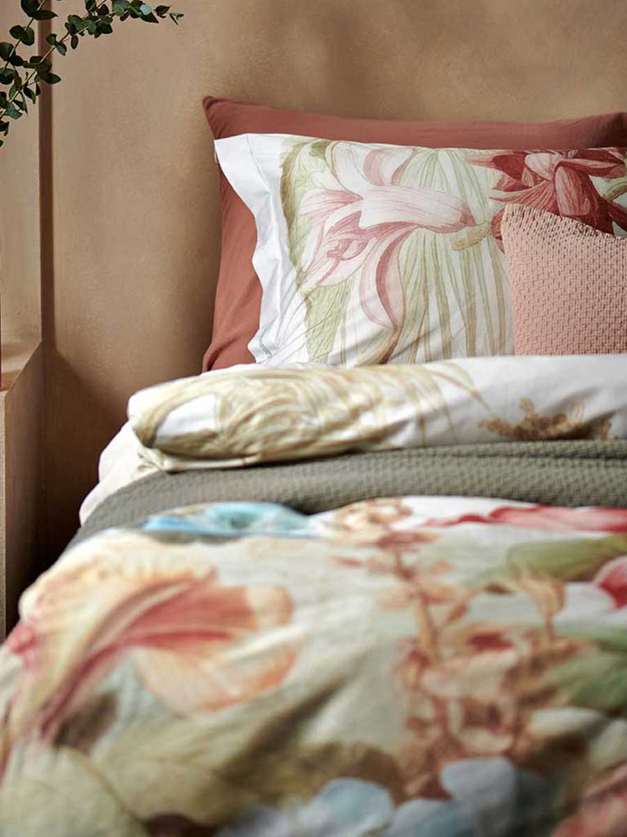 Dekbedovertrek met grote bloemenprint in warme tinten, gecombineerd met een terracotta hoeslaken en sierkussens, in een knusse slaapkamer met naturel muur.