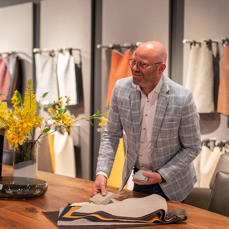 Interieurontwerper bij Home Center geeft persoonlijk designadvies aan een tafel met stofstalen en kleurmonsters, omgeven door een inspirerende presentatie van stoffen in diverse kleuren.