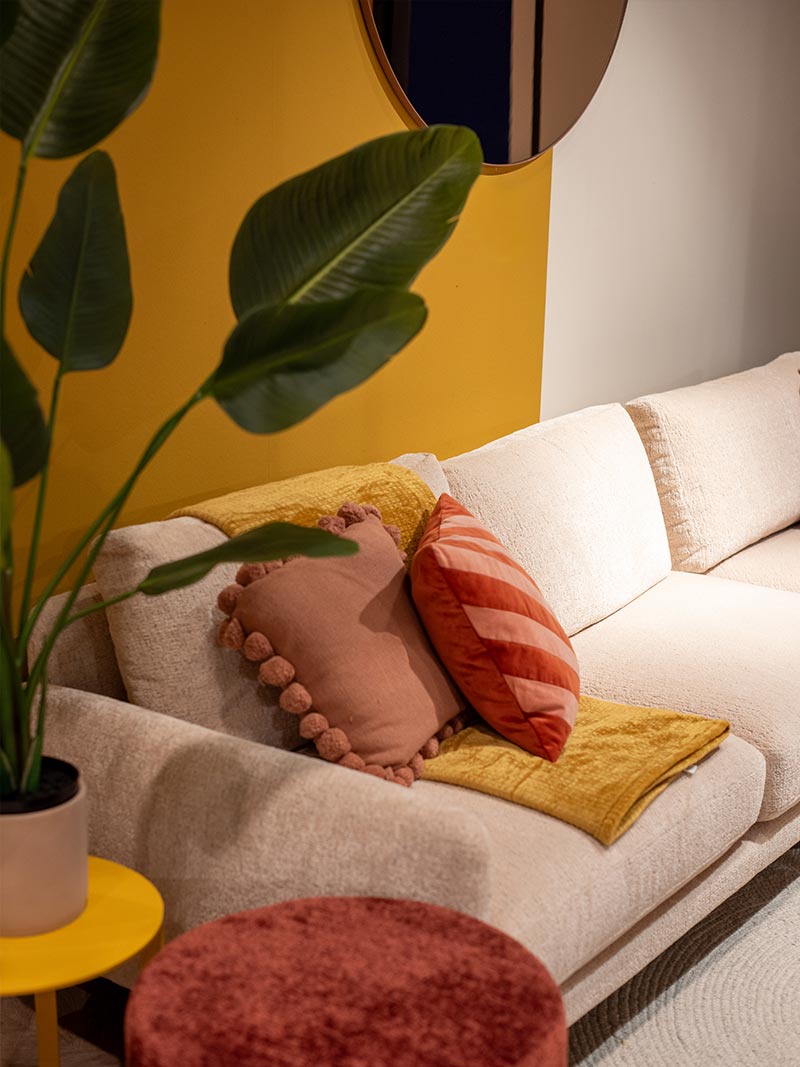 Beige hoekbank met roze en oranje kussens, gele plaid en grote groene kamerplant als eclectisch interieur