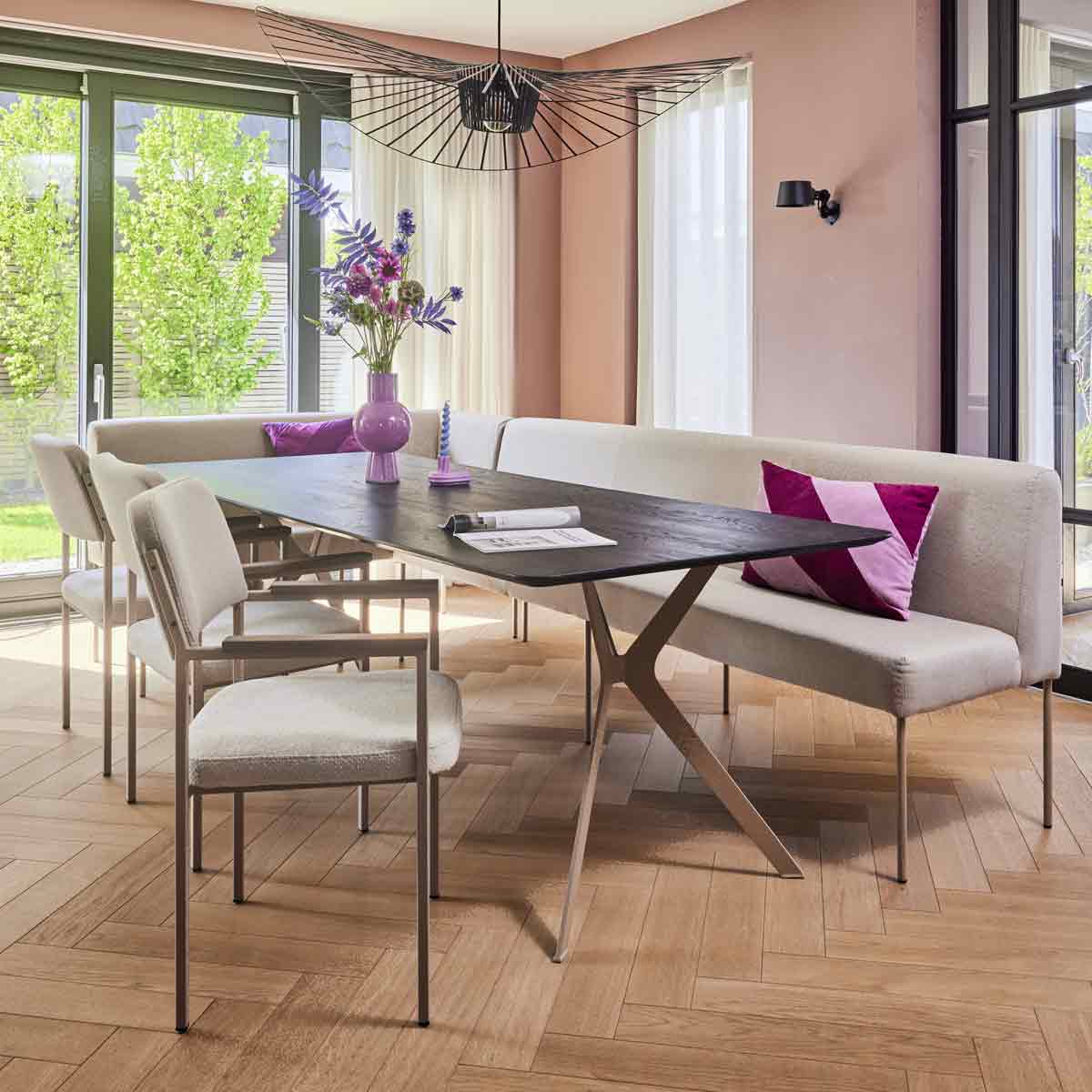 Een beige zitbank gecombineerd met beige eettafelstoelen aan een zwarte tafel met roze kussen