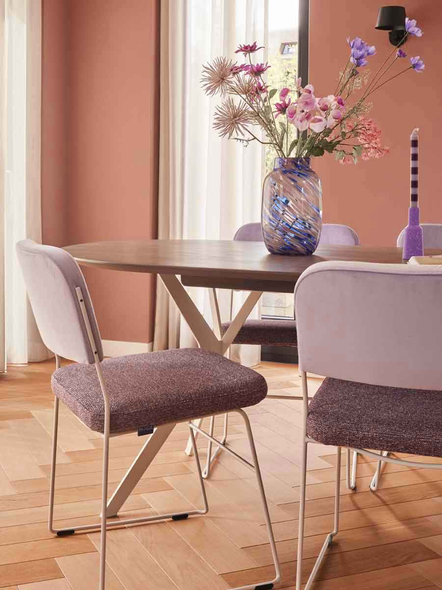 Twee velvet eetkamerstoelen met zwarte poten en verticale stiksels, in taupe en antraciet, voor een groot raam met uitzicht op een groene tuinmuur.