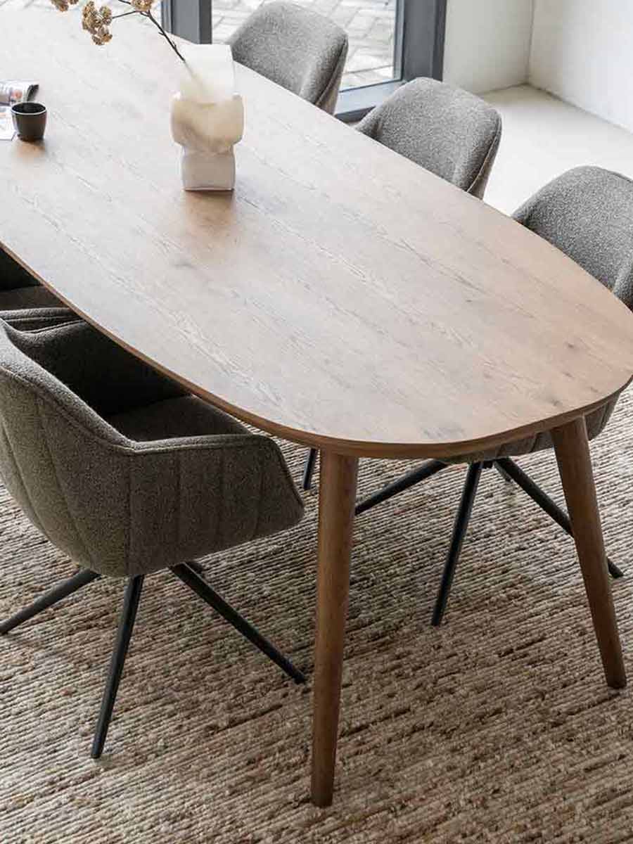 Houten ovale eettafel met naturel afwerking, omringd door draaibare designstoelen in zachte stoffen, op een geweven vloerkleed in een lichte eetkamer.