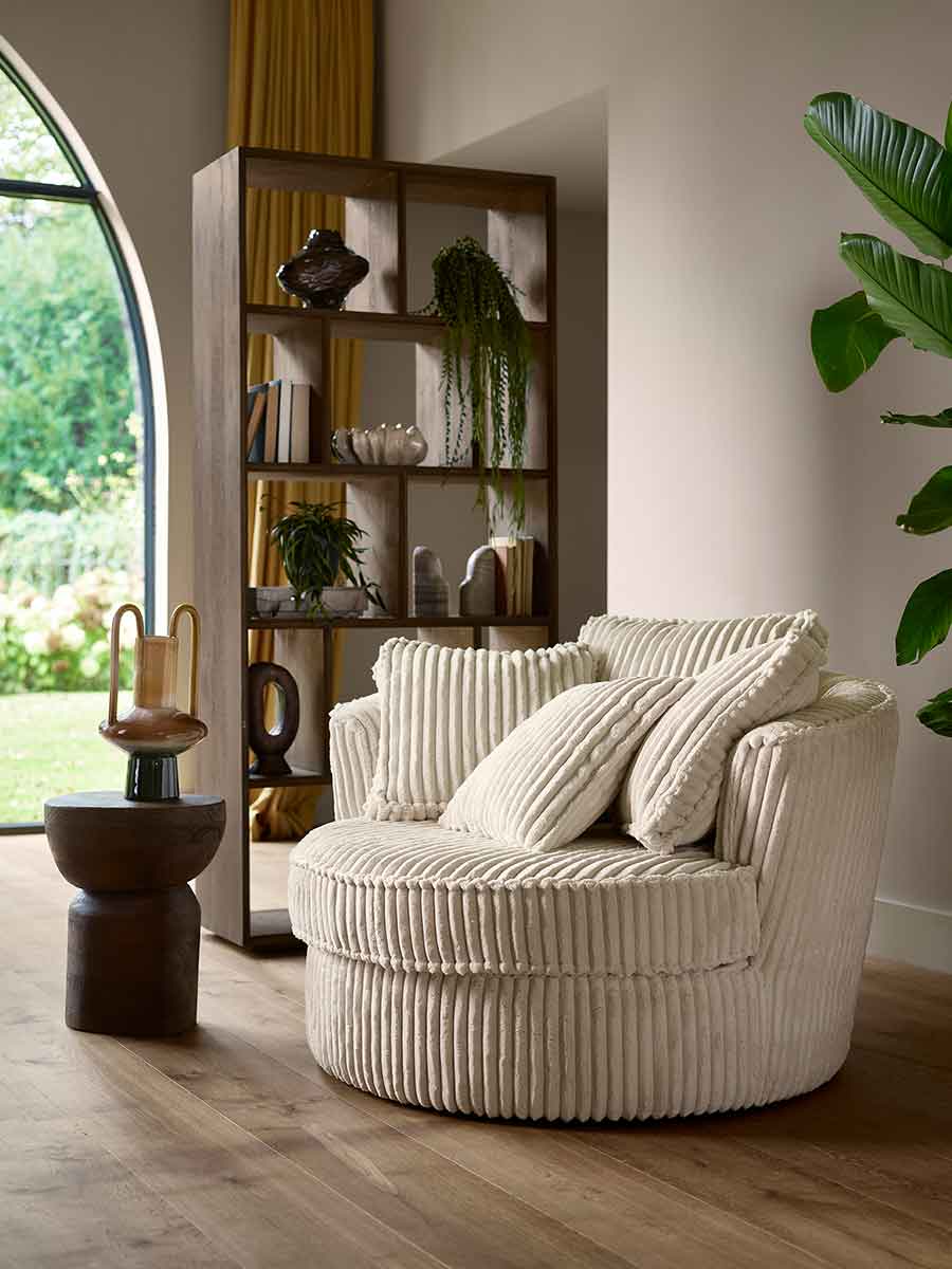 Crèmekleurige ronde fauteuil met ribstof bekleding en bijpassende sierkussens, geplaatst in een warme woonkamer met houten vloer en open wandkast op de achtergrond.