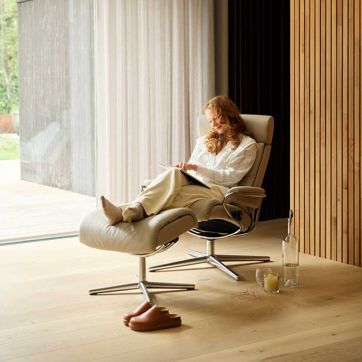 Vrouw ontspant in luxe relaxfauteuil met voetenbank bij groot raam en warme houten wand