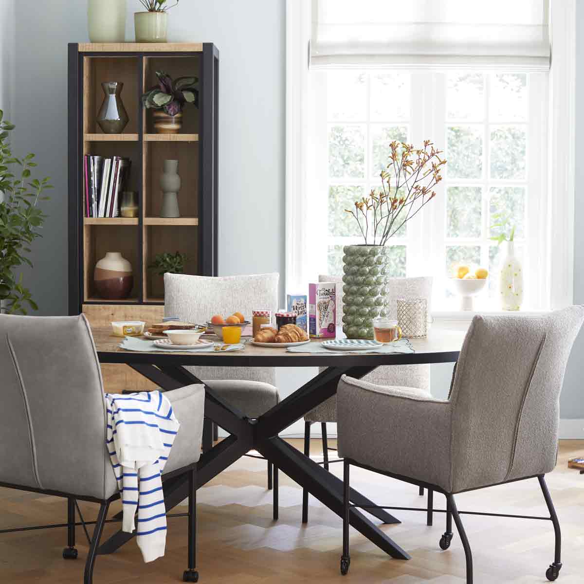 Moderne eetkamer met ovale houten tafel en gestoffeerde designstoelen in verschillende grijstinten. Een moeder en kind spelen samen aan tafel in een lichte ruimte met houten accenten.